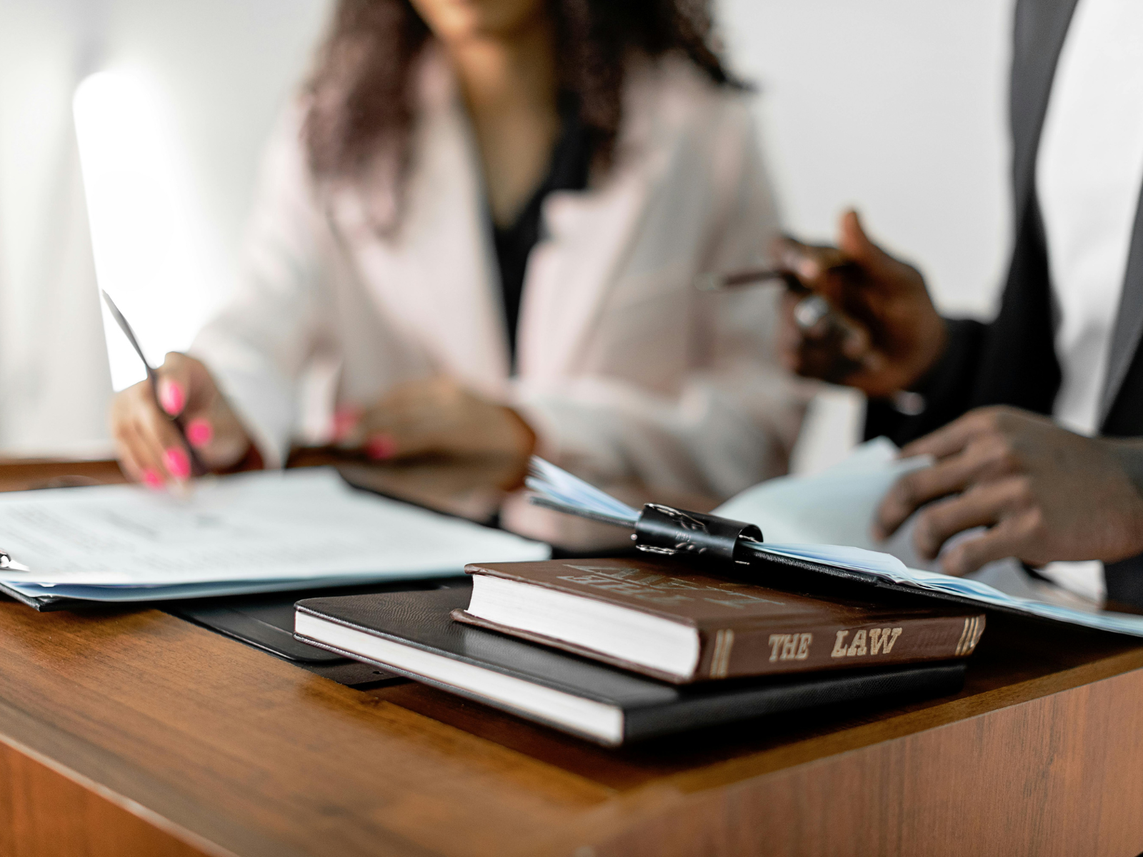 Close-up of a wooden desk with law books and documents, where two professionals are reviewing paperwork together. One person is wearing a light-colored suit and holding a pen, while the other wears a dark suit and holds a document.