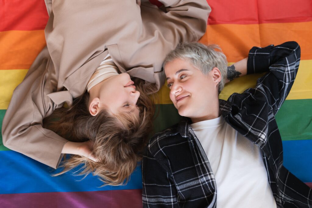 Young females lesbians LGBT couple lying on the rainbow flag looking at each other with love and passion for equality and sexual minorities rights. Transgender couple protesting for freedom in love. Estate planning for same-sex couples