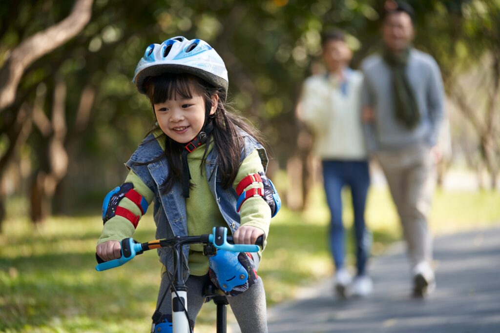 little asian girl with helmet and full protection gears riding bike in city park with parents watching from behind