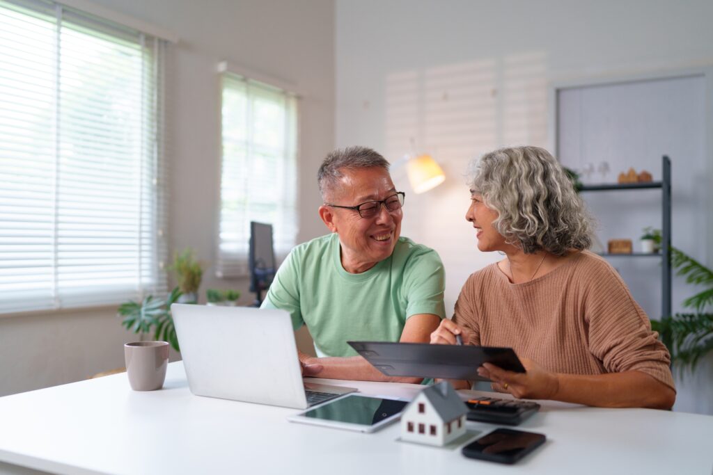 Happy older Asian couple managing finances and retirement planning at home. They use a laptop tablet and calculator while dreaming of a comfortable future together.